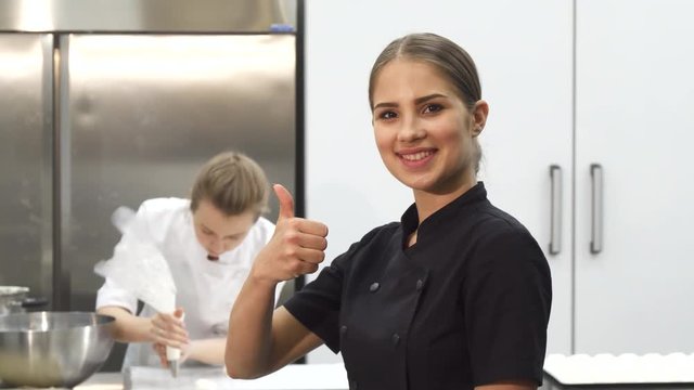 Stunning young beautiful female professional chef smiling joyfully to the camera showing thumbs up her colelague cooking at the kitchen on the background. Profession, employment.