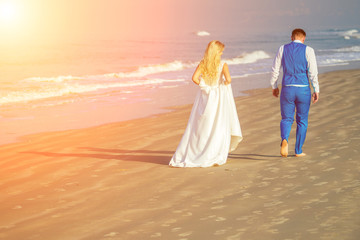 handsome groom in a chic suit and a beautiful bride in a wedding gown walk on the beach (view from the back). concept of a chic and rich wedding ceremony on the beach