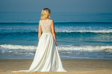 Fototapeta na wymiar young and beautiful bride with long blond hair standing his back and looking at the sea in a beautiful white long wedding dress. a happy bride married a wedding ceremony on the beach by the sea