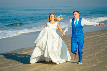 Fototapeta na wymiar handsome groom in a chic suit and a beautiful bride in a wedding dress are having fun and running on the beach. concept of a chic and rich wedding ceremony on the beach
