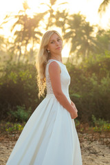 Fototapeta na wymiar charming and beautiful bride in a beautiful white long wedding dress with long blond hair smiling at the background of palm trees. happy bride married a wedding ceremony on the sea