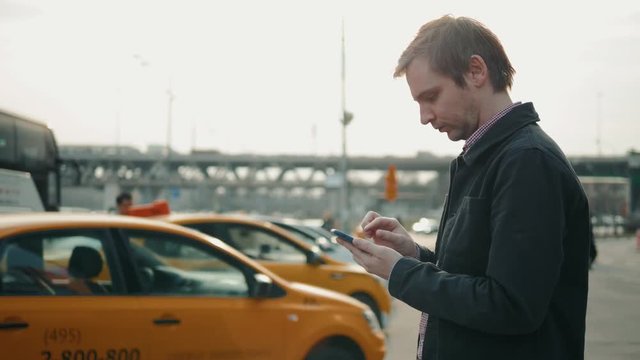 Elegant businessman checking taxi application app on mobile phone while walking outside airport, experienced male employer using cell telephone while waiting for trasportation orange taxi car outdoors