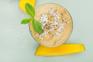 A glass of fruit smoothie on a blue wooden table with pieces of mango, with coconut shavings and a sprig of mint. Concept of healthy eating, top view, close-up.