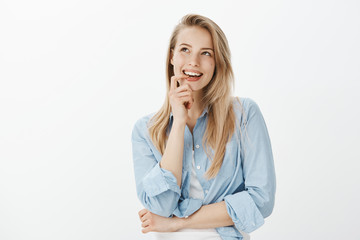 Creative good-looking female designer with blond hair, biting finger, looking up and smiling curiously while having great idea, standing dreamy over gray background, having interesting plan