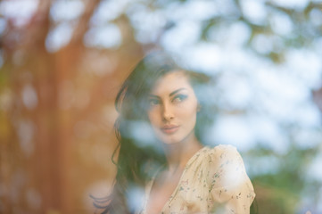 beautiful mysterious caucasian girl in summer dress behind window with reflections of trees