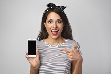 Naklejka premium Picture of glad fascinated young woman screaming in excitement, keeping mouth wide opened, feeling happy, demonstrating her new mobile phone, pointing index finger at blank copy space screen