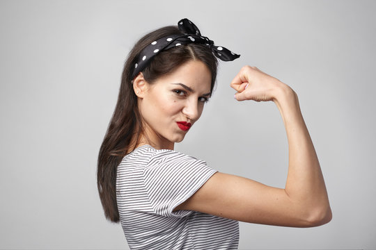 Picture of confident attractive young woman with dark loose hair and athletic slim body posing isolated at studio wall, demonstrating her strength and muscles, tensing bicep, staring at camera