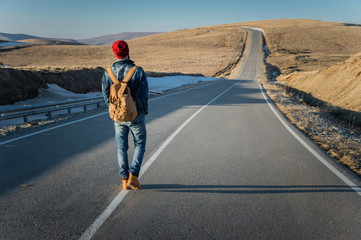 A bearded hipster with an old-fashioned vintage backpack wearing sunglasses with a red hat and jeans jacket and jeans walking goes with his back along a countryside asphalt mountain road in the