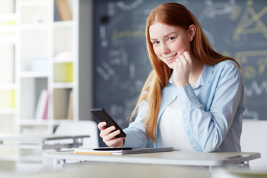 Smiling student looking at camera while sitting by her desk with smartphone and messaging at break
