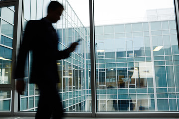 Fototapeta premium Blurred silhouette of mobile businessman walking along window inside modern office center