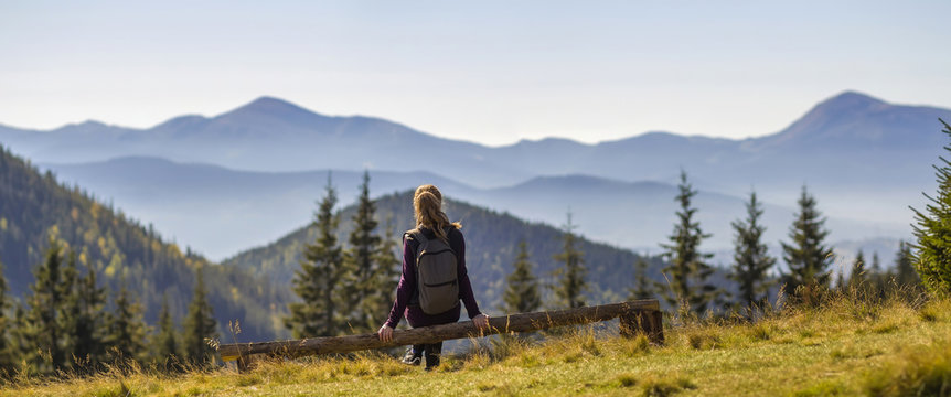 Back view of long-haired blond girl with backpack sitting on broken tree trunk enjoying breathtaking view of magnificent foggy Carpathian mountains, covered with ever-green forest on spring morning.