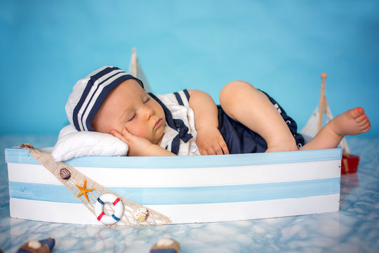 Cute toddler baby boy sleeping in wooden boat with fishes, starfish and binoculars, dressed as sailman