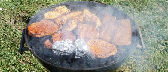 small round grill with pork and chicken and potato in aluminum foil over green grass in the garden