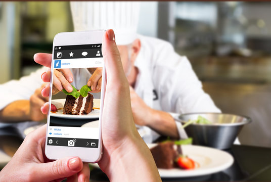 Hand holding smartphone against concentrated male pastry chef decorating dessert in kitchen