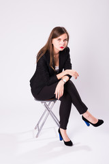 Fototapeta premium Full length studio portrait of young business woman in black suit, high heel shoes, sitting in chair on white background