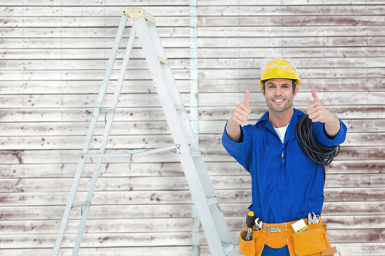 Happy electrician gesturing thumbs up by ladder against digitally generated grey wooden planks