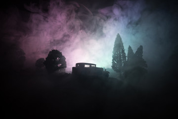 Silhouette of old vintage car in dark foggy toned background with glowing lights in low light, or silhouette of old car in dark forest.