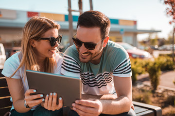 Young couple with sunglasses is sitting outside on a bench and using tablet