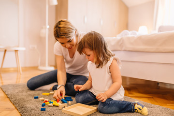Cute little girl is solving wooden puzzles with her mother on the floor in bedroom
