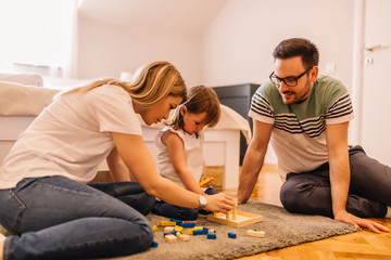Young parents are helping to their daughter with solving puzzles on the floor in bedroom