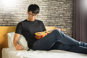 Happy young asian man playing ukulele sitting on bed in bedroom. - Powered by Adobe