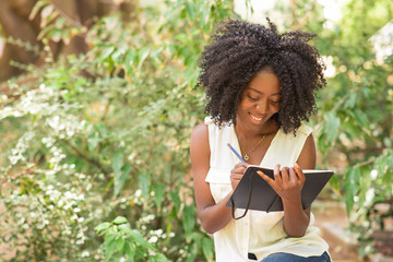 Happy attractive young black woman making notes in park. Girl writing and sitting with plants in...
