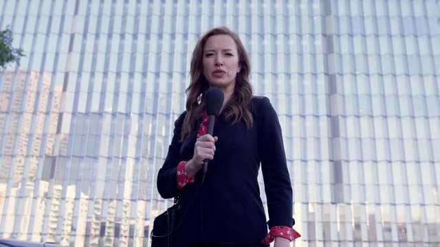 MS smartly dressed female TV journalist talks with microphone in front of windows of new Federal Court building in Downtown Los Angeles. Hand-held, real time 4K UHD. Mute