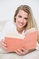 Smiling gorgeous model lying on cosy bed reading book