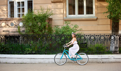 Fototapeta premium Lovely woman on a vintage bicycle is riding along the city street looking at the camera. The girl is dressed in a light dress and an accessory on her head