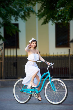 Joyful young attractive woman wearing white dress enjoying riding bicycle in the city smiling to the camera transportation vehicle tourism tourist urban lifestyle seasonal concept.