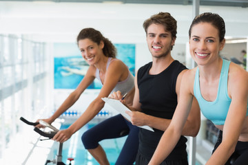 Obraz premium Fit women in a spin class with trainer taking notes and smiling at camera