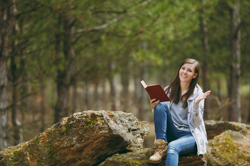 Young smiling beautiful woman sitting on stone studying reading book and spreading hands in city park or forest on green blurred background. Student learning, education. Lifestyle, leisure concept.