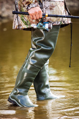 fisherman stands in rubber boots for a swamp in the river and holds a fishing rod in his hand in the summer