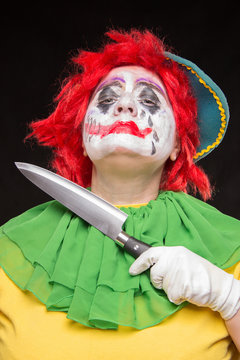 Scary clown with a horrible make-up laughs and with knives in hands on a black background