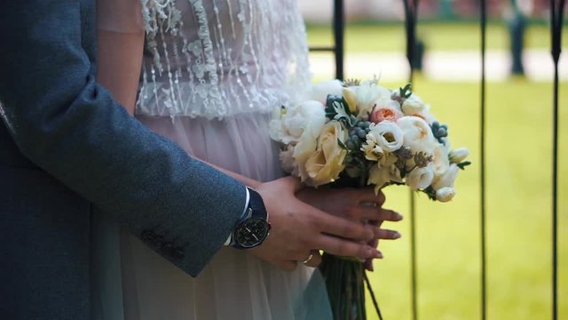 The groom puts his hands on the abdomen of the bride. The bride holds flowers in her hands. Close-up only hands