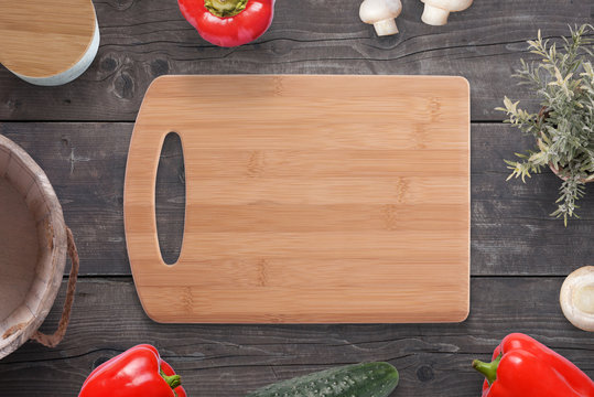 Empty cutting board on wooden desk surrounded with vegetables, mushrooms, spice box, plant, milk bucket.