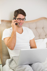 Casual smiling man using cellphone and laptop in bed