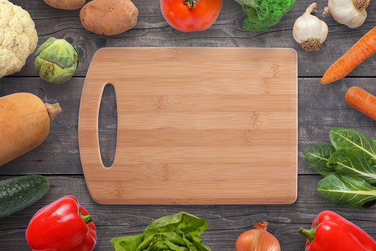 Food cutting board surrounded with vegetables on black wooden desk. Vintage, flat lay, top view.