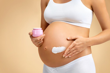 Close up of pregnant woman applying moisturizing cream on her belly on beige background. Pregnancy, maternity, preparation and expectation concept - obrazy, fototapety, plakaty