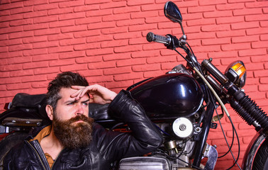 Fototapeta na wymiar Bikers lifestyle concept. Hipster, brutal biker on pensive face in leather jacket sits, leans on motorcycle. Man with beard, biker in leather jacket near motor bike in garage, brick wall background.