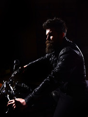 Fototapeta na wymiar Man with beard, biker in leather jacket sitting on motor bike in darkness, black background. Hipster, brutal biker in leather jacket riding motorcycle at night time, copy space. Night rider concept.