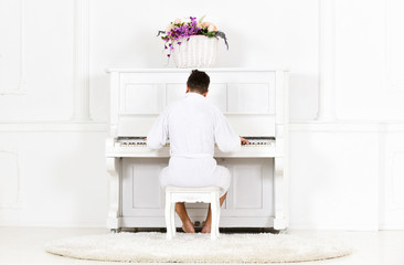 Man in bathrobe enjoys morning while playing piano. Talented musician concept. Man sleepy in bathrobe sit in front of piano musical instrument in white interior on background, rear view.