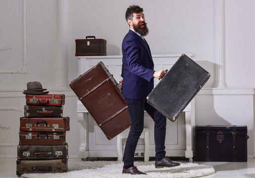 Man, traveller with beard and mustache with luggage, luxury white interior background. Macho elegant on cheerful face walks near pile of vintage suitcase, holds suitcases. Hotel service concept.
