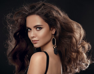 Glamour brunette. Fashion portrait of gorgeous sexy woman with long healthy hair style and evening makeup, black earrings jewelry looking at camera isolated on black studio background with back light.
