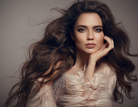Beauty fashion brunette portrait. Sexy woman wears in stylish fur coat. Beautiful girl model with evening makeup, long healthy curly hair style posing over studio gray background.