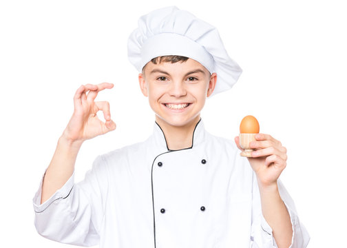 Handsome teen boy wearing chef uniform holding brown egg. Portrait of a happy cute male child cook making ok gesture, isolated on white background. Food and cooking concept.