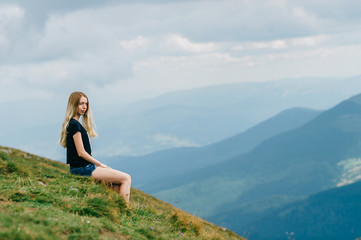 Naklejka na ściany i meble Young girl relaxing and enjoying scenic view on top of mountain. Bliss and happiness. Beautiful summer nature landscape. Lonely young blonde teen traveler dreaming outdoor on vacation. Romantic mood
