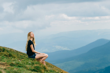 Naklejka premium Young girl relaxing and enjoying scenic view on top of mountain. Bliss and happiness. Beautiful summer nature landscape. Lonely young blonde teen traveler dreaming outdoor on vacation. Romantic mood