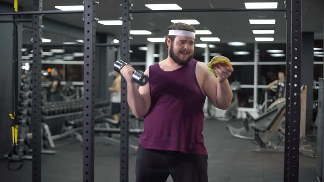 Obese man holding dumbbell eating burger, refusing from doing sport, addiction