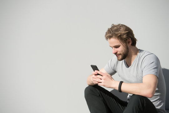 Bearded man use mobile phone on grey wall. Macho with smartphone on sunny outdoor. Guy with beard and blond hair in casual tshirt text sms. New technology and modern life, copy space